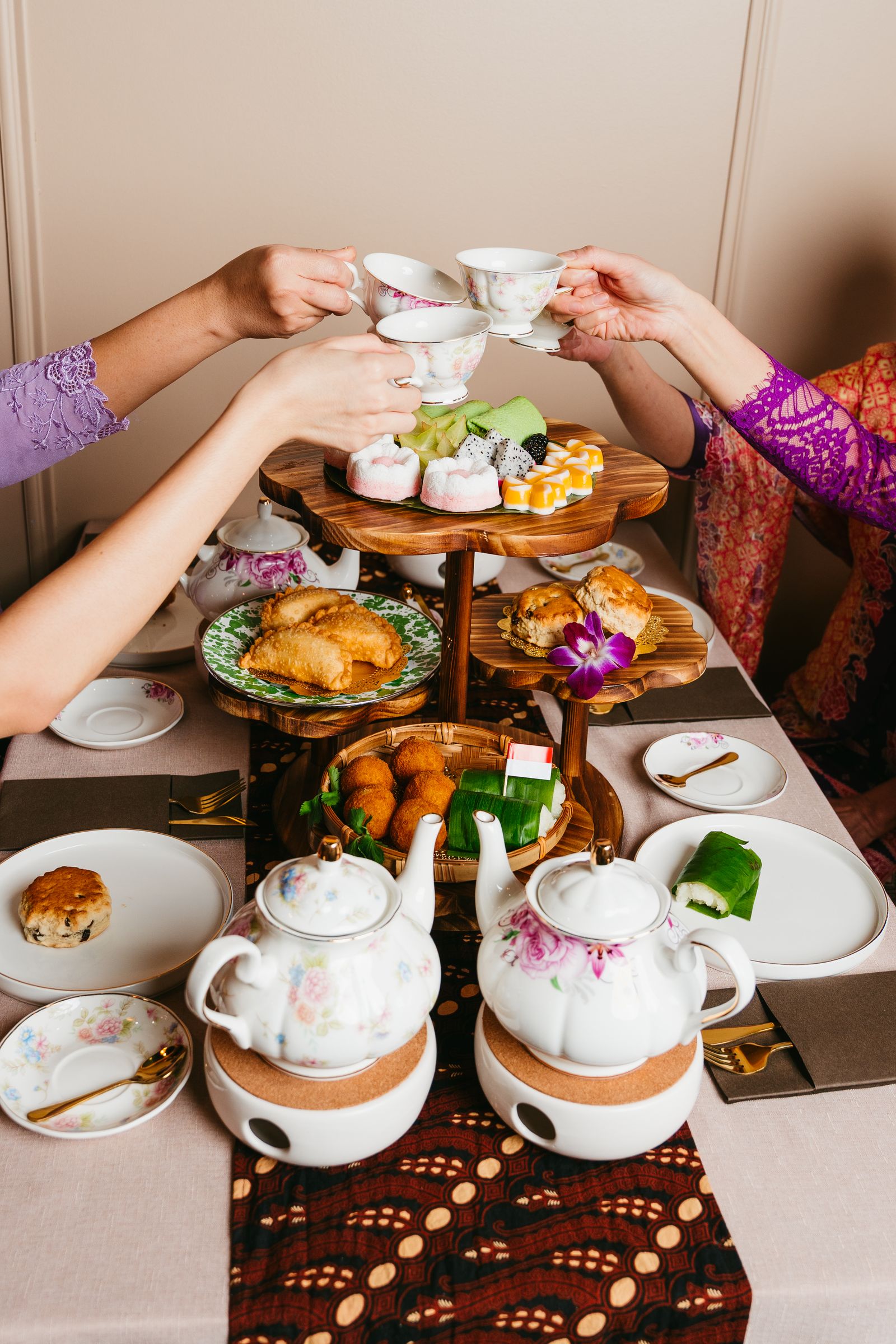 Two guests toasting teacups over a tiered spread of Indonesian desserts and tea at gula