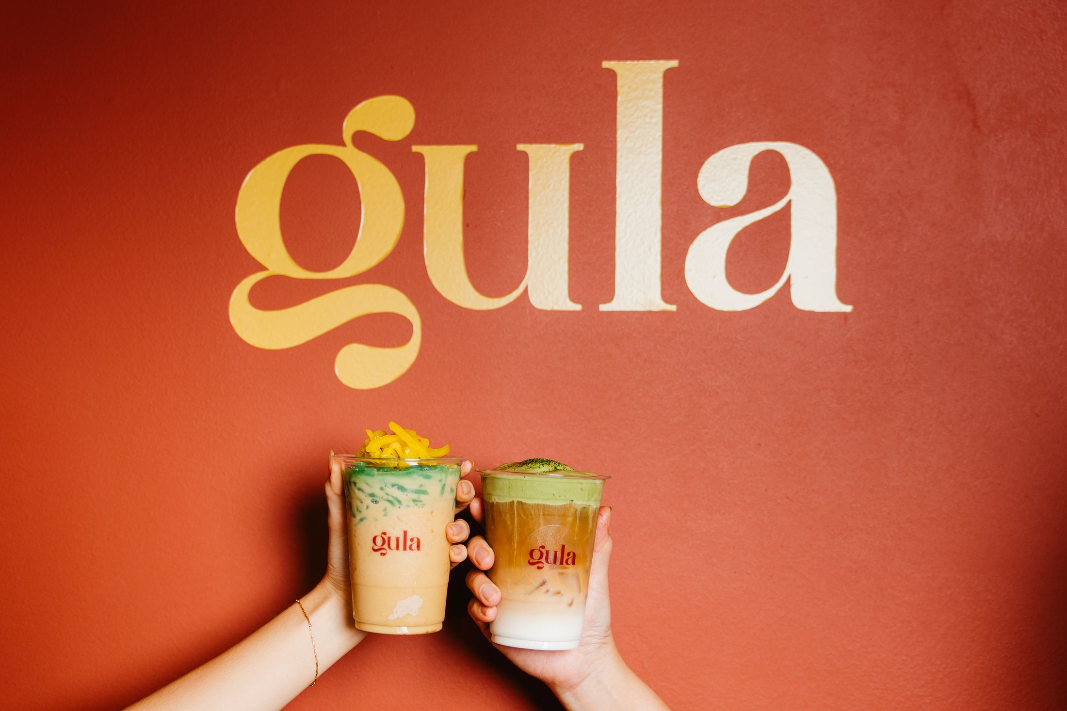 Two hands toasting iced gula drinks in front of the golden gula wordmark on a terracotta wall
